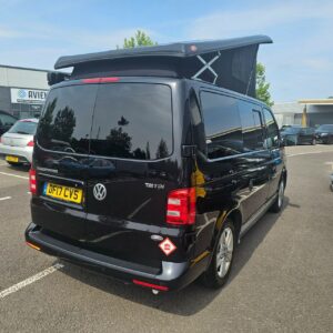 A SWB T6.1 Volkswagen Transporter Highline Campervan in Deep Black (17 Plate, A1466) is parked in a lot with trees and buildings in the background.