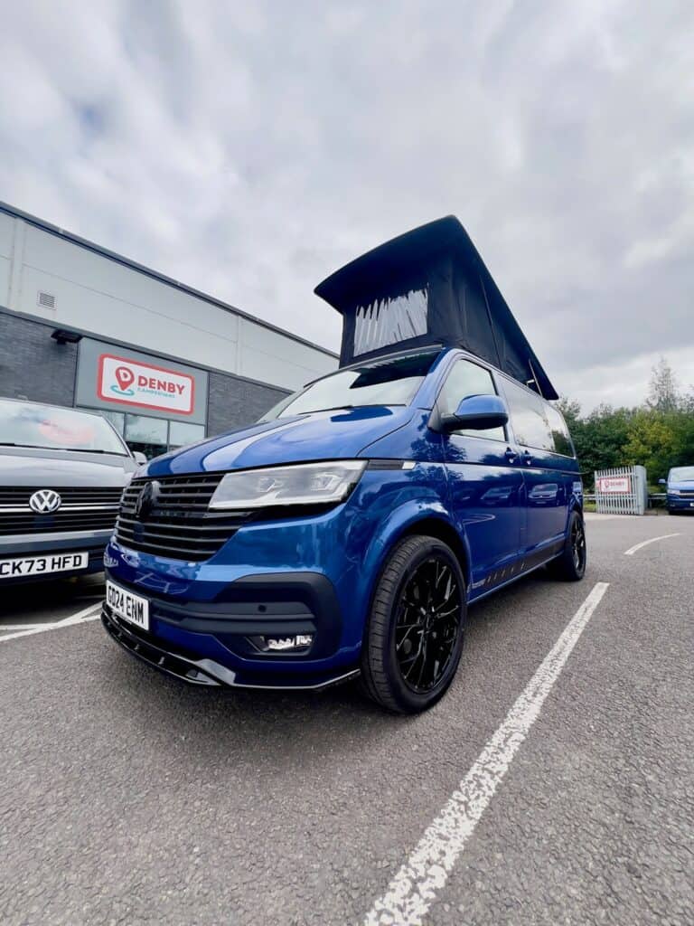 A Ravenna Blue SWB T6.1 Volkswagen Transporter Highline Campervan (Automatic, 24 plate, A1471) is parked outside a building with a Denby sign next to another Volkswagen vehicle under a cloudy sky.