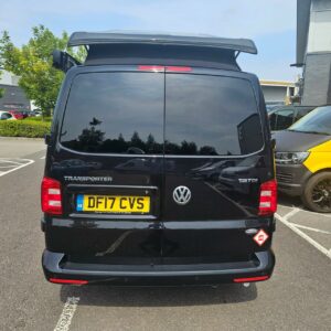 A Deep Black SWB T6.1 Volkswagen Transporter Highline Campervan (17 Plate, A1466) is parked in a lot, its rear windshield reflecting trees and buildings. The partially raised roof and a sticker on the lower right are visible.