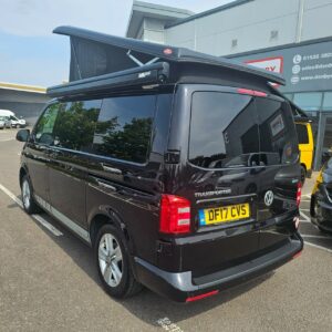 A SWB T6.1 Volkswagen Transporter Highline Campervan, Deep Black, 17 Plate (A1466), with a pop-top roof is parked outside a commercial building, its windows reflecting trees.