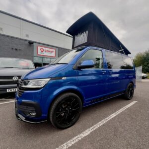 A Ravenna Blue Volkswagen T6.1 Transporter Highline SWB campervan (Automatic, 24 plate, A1471) with a raised pop-top roof is parked near a Denby-signed building on an overcast day, with another car nearby.