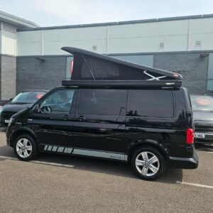 A SWB T6.1 Volkswagen Transporter Highline Campervan in Deep Black (17 Plate, A1466) is parked in a lot among other vehicles, in front of a modern building with grey walls and large windows.