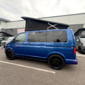 A Ravenna Blue SWB T6.1 Volkswagen Transporter Highline Campervan (24 plate, A1471, automatic) with a pop-top roof is parked outside a grey industrial building among other vans under an overcast sky.