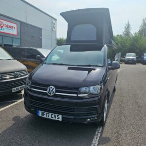 A SWB T6.1 Volkswagen Transporter Highline Campervan in Deep Black (17 Plate, A1466) is parked outside a building with Denby Campervans signage, with other vehicles and trees in the background.