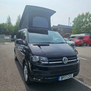 A SWB T6.1 Volkswagen Transporter Highline Campervan in Deep Black (17 Plate, A1466) is parked in a lot on a sunny day, with other vehicles and buildings visible in the background.