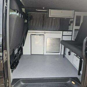 The image shows the interior of a SWB T6.1 Volkswagen Transporter Highline Campervan (Deep Black, 17 Plate, A1466) with folded seats, a compact kitchenette (fridge, oven, cabinets, drawer), grey curtains on the window, and clean grey flooring.