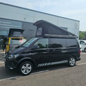 A SWB T6.1 Volkswagen Transporter Highline Campervan in Deep Black (17 Plate, A1466) with a raised pop-top roof is parked outside a building, with another yellow campervan and the building visible in the background.