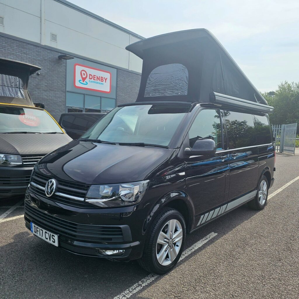 A SWB T6.1 Volkswagen Transporter Highline Campervan in Deep Black (17 Plate, A1466) with a pop-up roof is parked outside a building with a Denby sign, next to another vehicle on a sunny day.