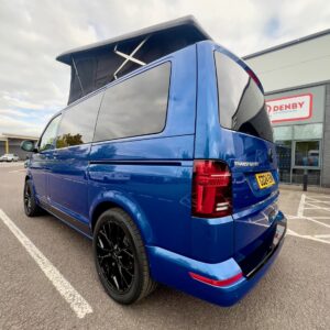 A Ravenna Blue SWB T6.1 Volkswagen Transporter Highline Campervan (24 plate, A1471) with black wheels and an extended pop-up roof is parked outside a building with a DENBY sign under partly cloudy skies.