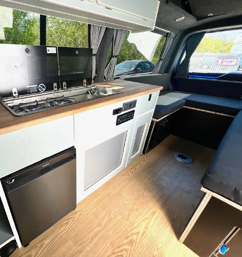 Modern camper van interior with a compact kitchen featuring a stove, sink, mini fridge, light cabinetry, and a seating area with cushions. Sunlight streams in through large windows with curtains.