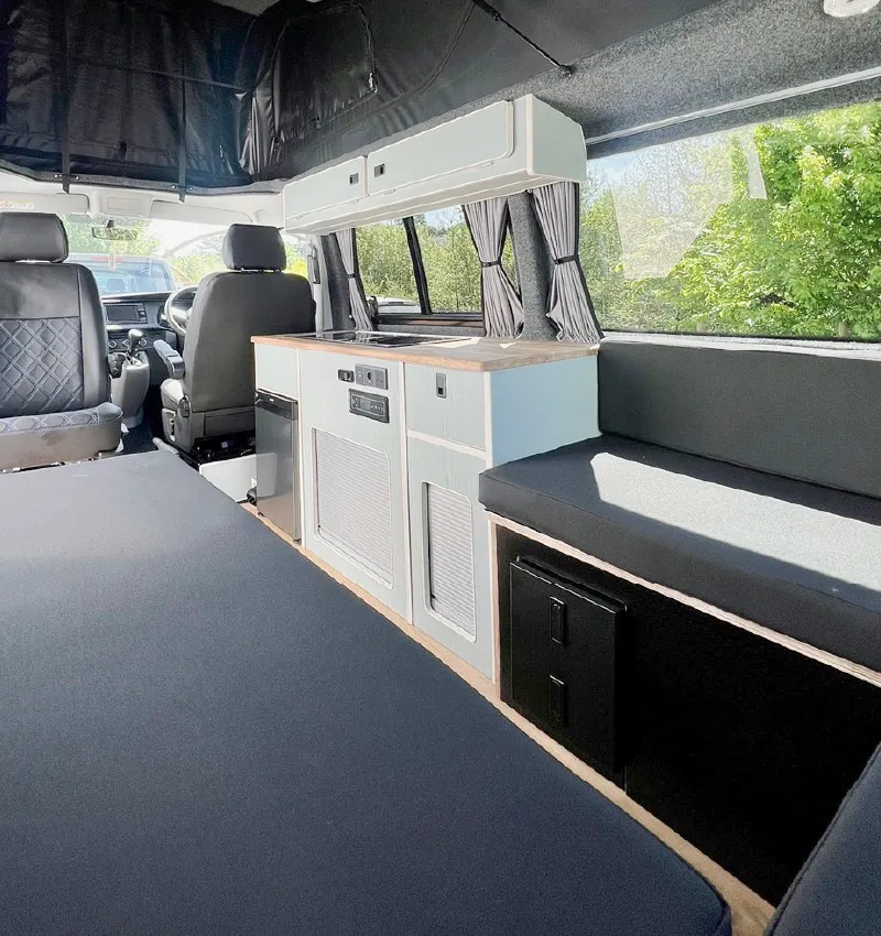 Interior of a camper van with a small kitchenette, black cushioned seating, storage cabinets, and large windows with gray curtains letting in sunlight. The drivers area is visible in the background.