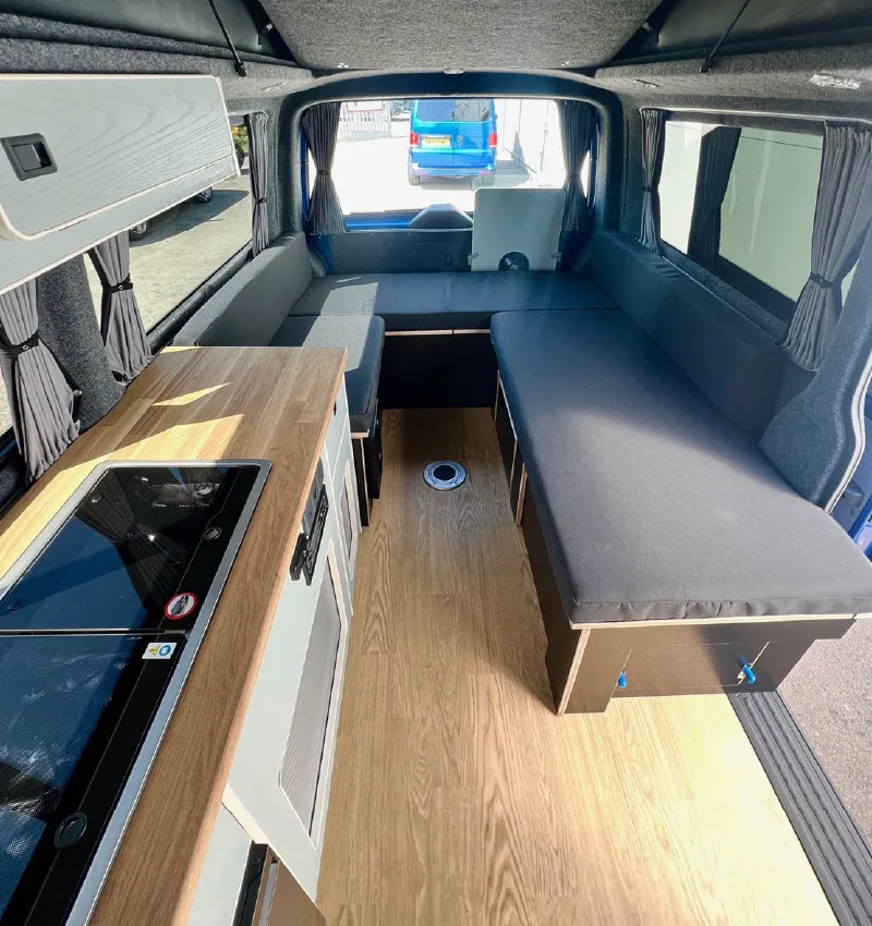 Interior of a camper van with a wooden floor, a compact kitchenette on the left, black cushioned seating that can convert into a bed, and windows with gray curtains on both sides.