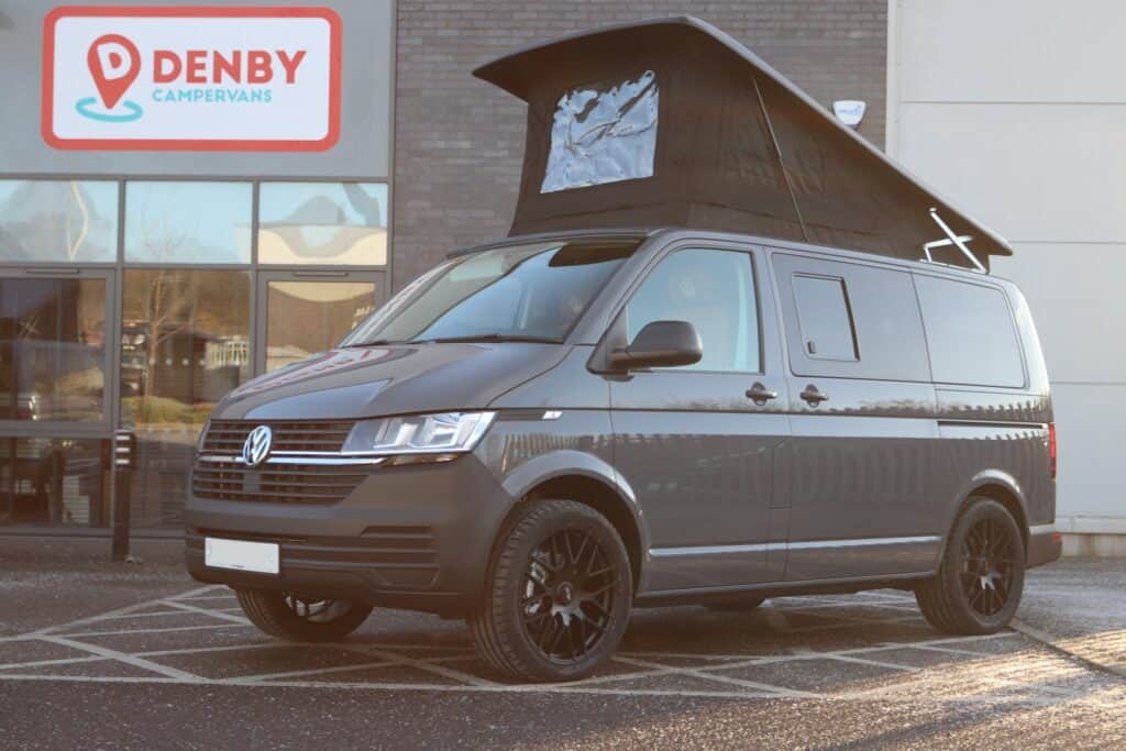 A 21 plate Balmoral Deluxe SWB - T6.1 Volkswagen Transporter Startline Campervan in Pure Grey with a raised pop-top roof is parked outside a building displaying a Denby Campervans sign, showing both its front and side.