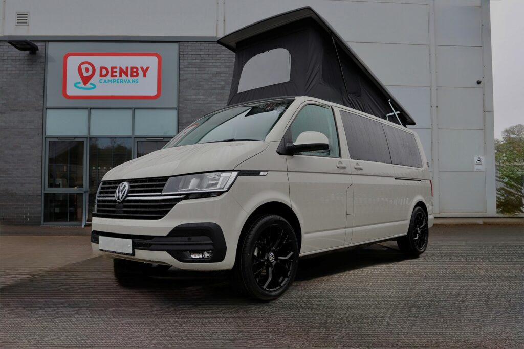 An Ascot Grey Volkswagen Transporter T6.1 LWB campervan (150BHP Automatic, 22 Plate, A1481) is parked in front of a building with a Denby Campervans sign.