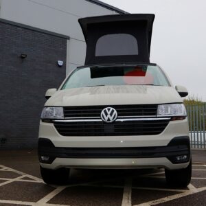 An Ascot Grey LWB T6.1 Volkswagen Transporter Campervan (150BHP-Automatic, 22 Plate, A1481) is parked with its pop-up roof raised in a disabled bay outside a modern building on a cloudy day.
