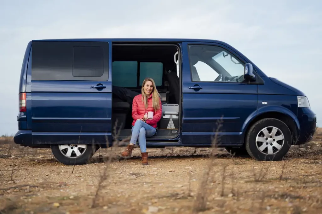 A woman in a red jacket sits smiling in the open side door of a blue van parked on a dry, grassy field under a cloudy sky.