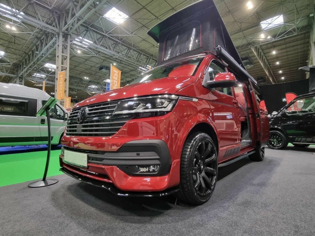 The Balmoral Deluxe SWB - T6.1 Volkswagen Transporter Highline Campervan in Fortana Red (24 Plate, A1485) is displayed indoors at an exhibition, featuring a pop-up roof, black wheels, and open side door under bright lights.
