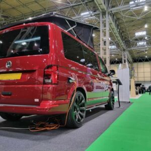 The Balmoral Deluxe SWB - T6.1 Volkswagen Transporter Highline Campervan in Fortana Red (24 Plate, A1485) is displayed indoors at an exhibition, with orange cables plugged near the rear wheel and a green carpeted aisle in the background.
