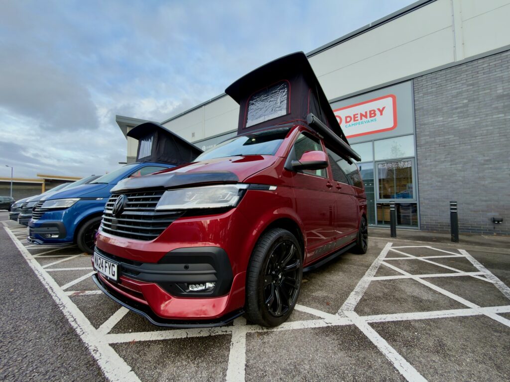 A Fortana Red Balmoral Deluxe SWB T6.1 Volkswagen Transporter Highline Campervan (24 Plate, A1485) with a raised pop-up roof is parked in a marked bay outside Denby Campers, next to a blue van.