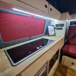 The Balmoral Deluxe SWB T6.1 VW Transporter Highline Campervan (Fortana Red, 24 Plate – A1485) features a modern interior with red quilted seats, wood countertop, built-in stovetop, red blinds, control panels, and framed decor.