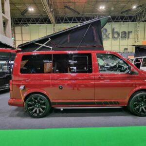 A Balmoral Deluxe SWB - T6.1 Volkswagen Transporter Highline Campervan in Fortana Red (24 Plate, A1485) with black wheels and detailing is parked indoors at an exhibition, with other vehicles and stands in the background.