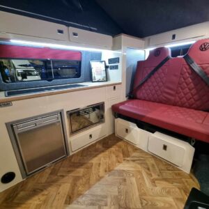 The Balmoral Deluxe SWB T6.1 Volkswagen Transporter Highline Campervan (Fortana Red, 24 Plate – A1485) features a modern interior with red leather seats, wood-patterned floor, compact kitchen with fridge, cabinets, microwave, storage under seats and soft overhead lighting.